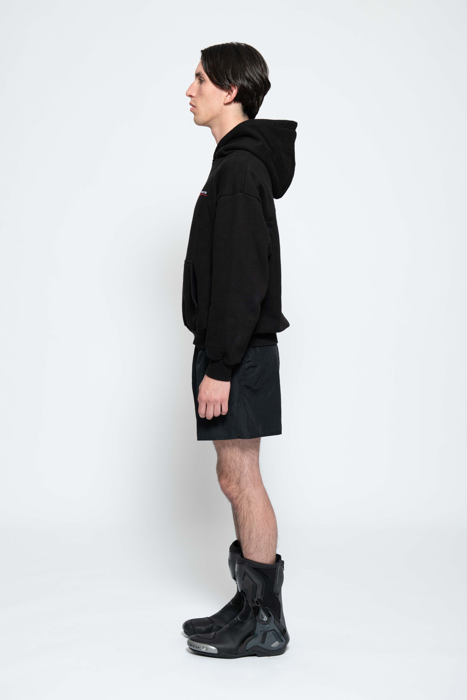 Seitenansicht, Ganzkörperbild. Männliches Model, mit einem schwarzen Unisex "Swoosh Zements" Hoodie. Dazu trägt das Model eine kurze schwarze Short und schwarze Bikerstiefel