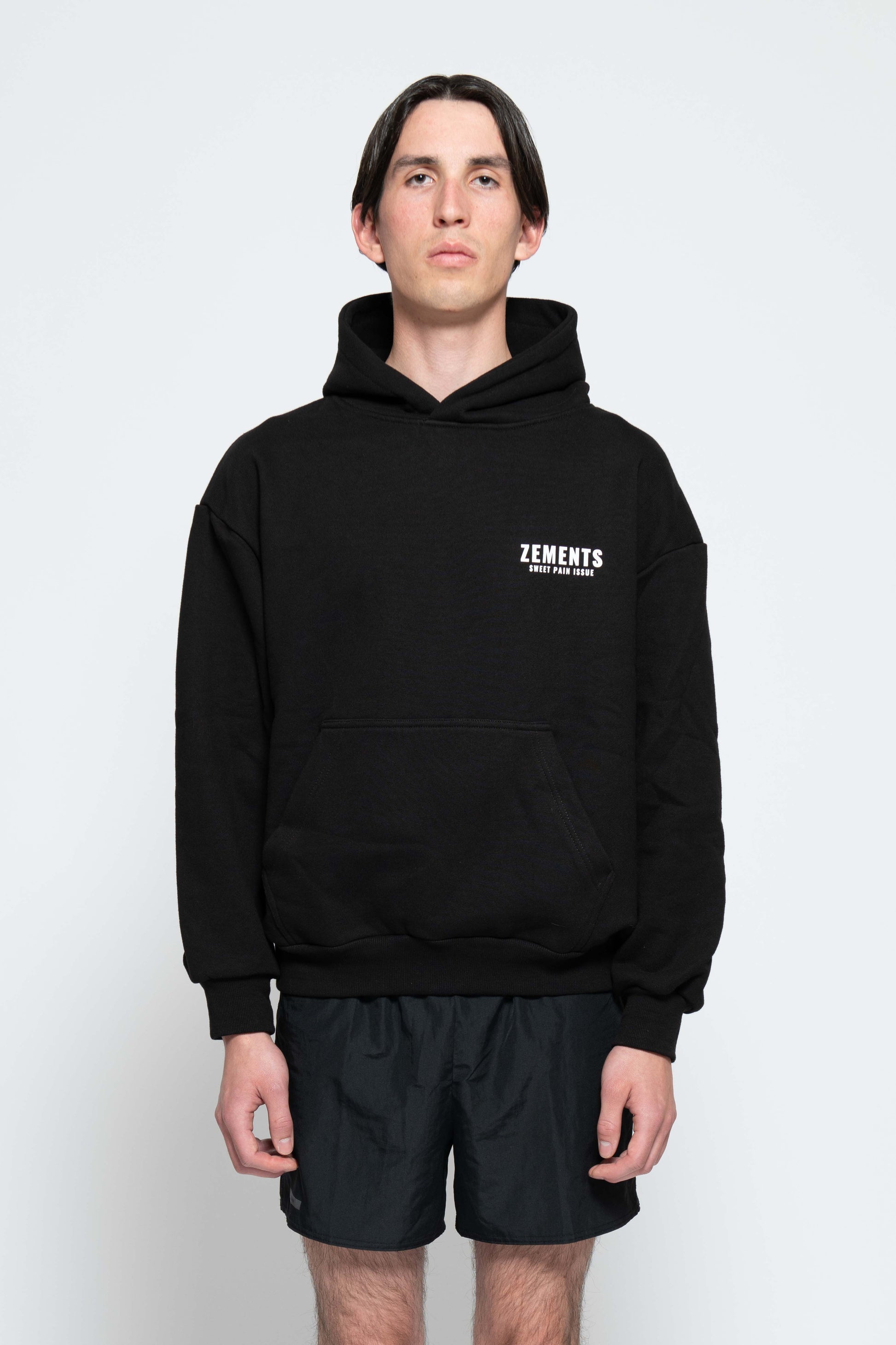 Frontalansicht, Halbkörperbild. Männliches Model, mit einem schwarzen Unisex„Sweet Pain Issue“ Hoodie. Oben Links auf Brust-Höhe, ist das „Zements-Logo„ und drunter das „Sweet Pain Issue-Logo“ in Weiß klein aufgedruckt.
