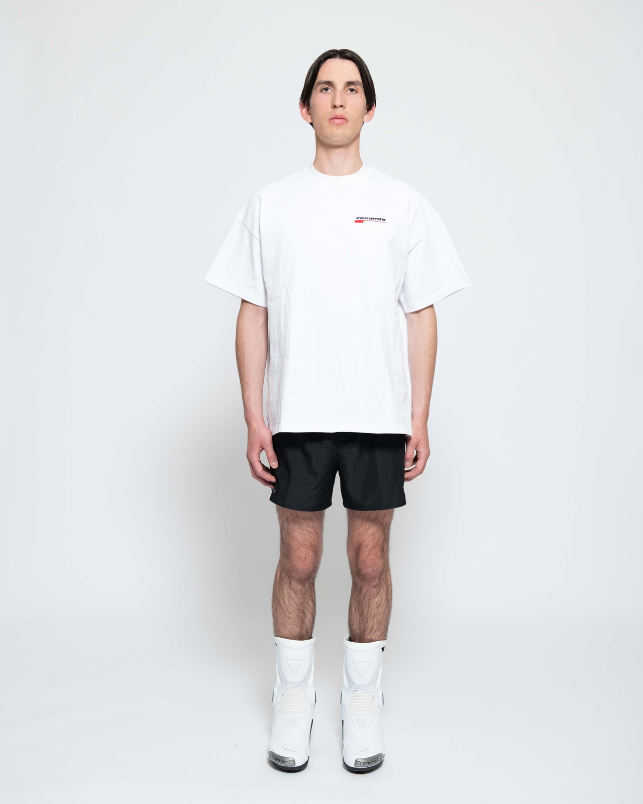 Frontalansicht, Ganzkörperbild. Männliches Model, mit einem weißen oversized "Drift Zements" T-shirt. Oben Links auf Brust-Höhe, ist das "Swoosh Zements-Logo" in Rot/Schwarz klein aufgestickt. Dazu trägt das Model eine kurze schwarze Short und schwarze Bikerstiefel