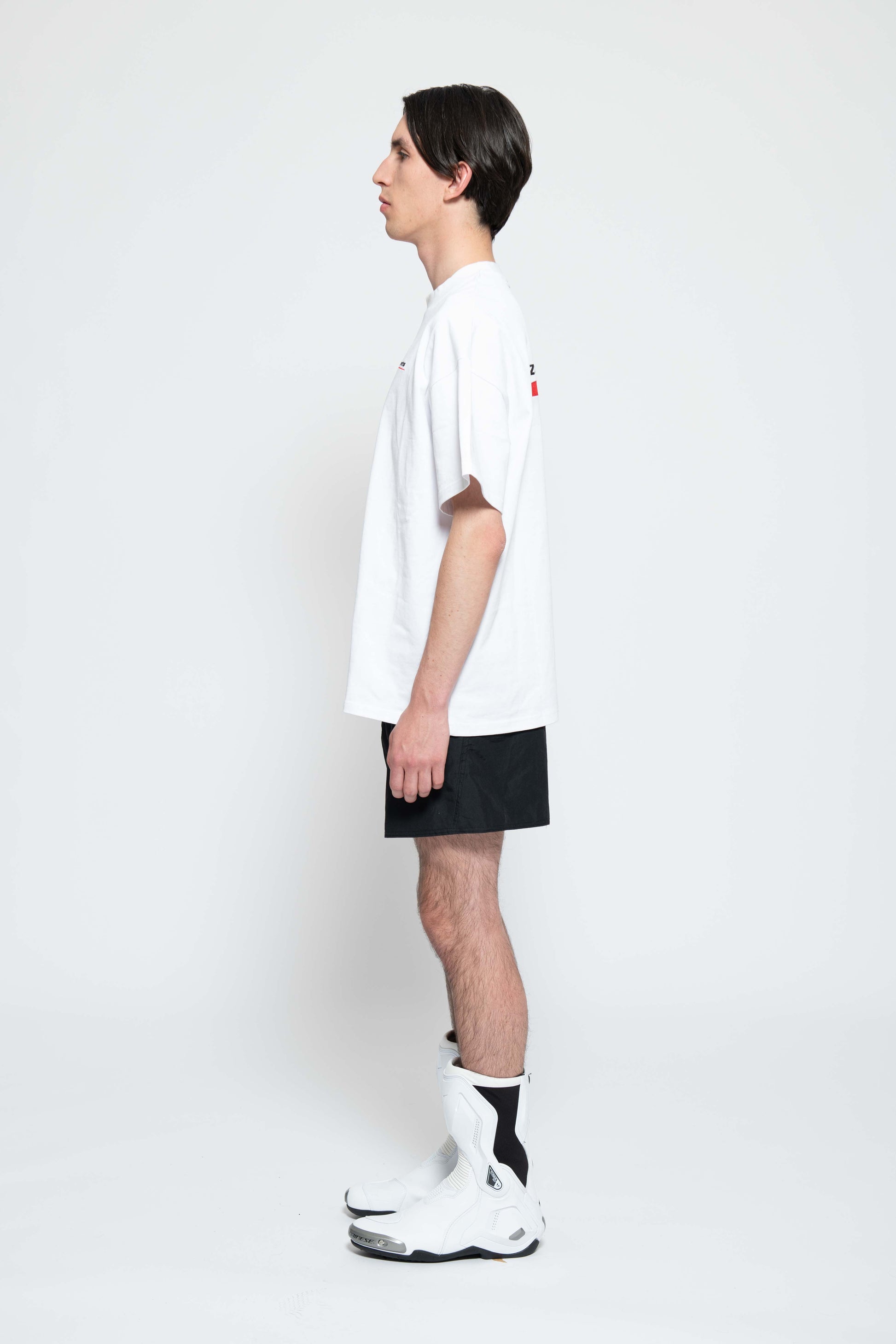 Seitenansicht, Ganzkörperbild. Männliches Model, mit einem weißen oversized "Swoosh Zements" T-shirt. Dazu trägt das Model eine kurze schwarze Short und schwarze Bikerstiefel