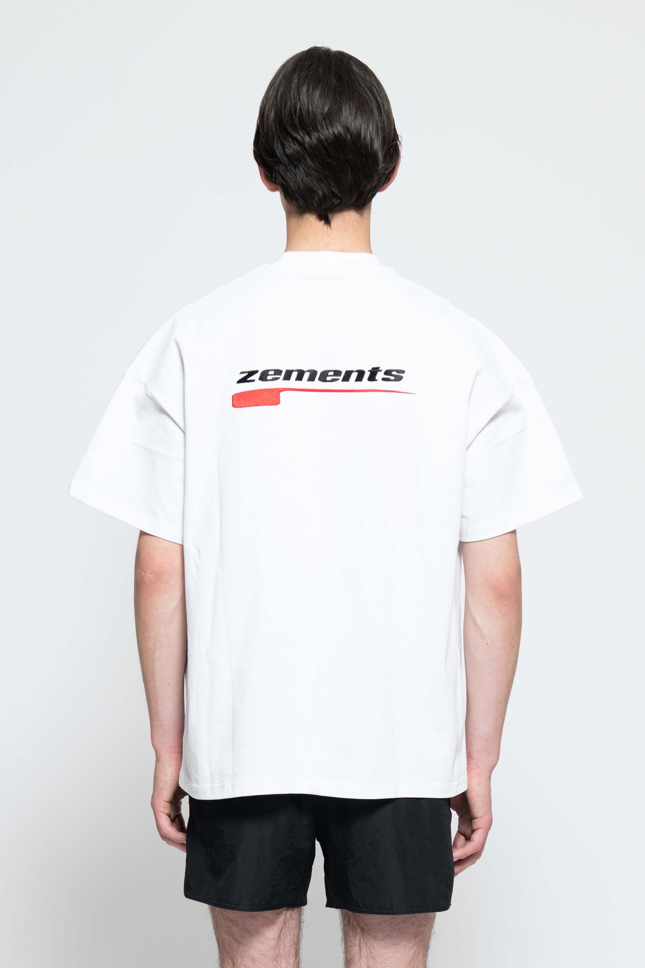Frontalansicht, Ganzkörperbild. Männliches Model, mit einem weißen oversized "Drift Zements" T-shirt. Oben Links auf Brust-Höhe, ist das "Swoosh Zements-Logo" in Rot/Schwarz klein aufgestickt. Dazu trägt das Model eine kurze schwarze Short und schwarze Bikerstiefel